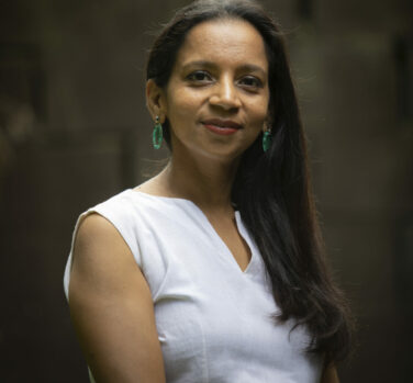 A young Indian woman with long hair across one shoulder looks at the camera and smiles, lips closed