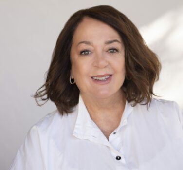 A middle-aged woman with shoulder-length brown hair and a white button up shirt smiles at the camera