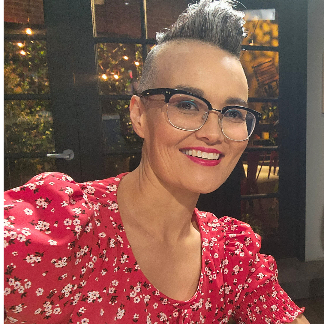 A woman with a short gray mohawk and dark-rimmed glasses leans into the frame, smiling