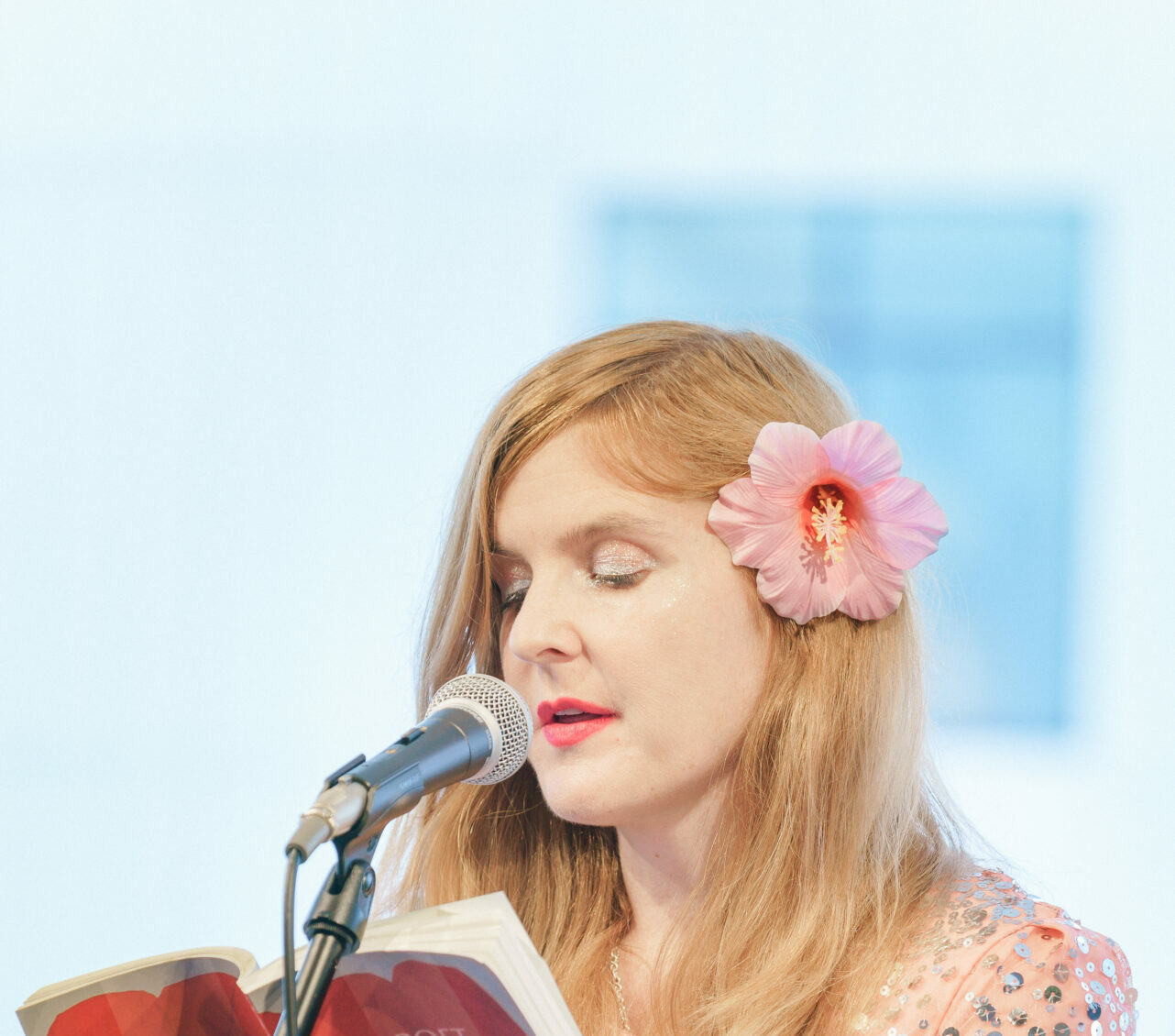 A blonde woman with a pink flower in her hair reads from a book at a microphone
