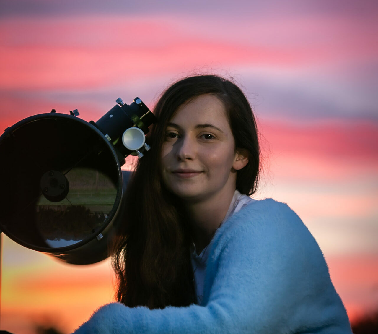 A young woman with long brown hair sits beside a telescope, the sky behind her is purple and pink