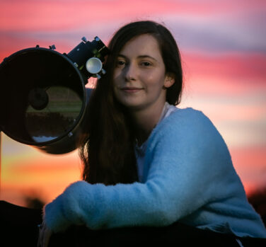 A young woman with long brown hair sits beside a telescope, the sky behind her is purple and pink