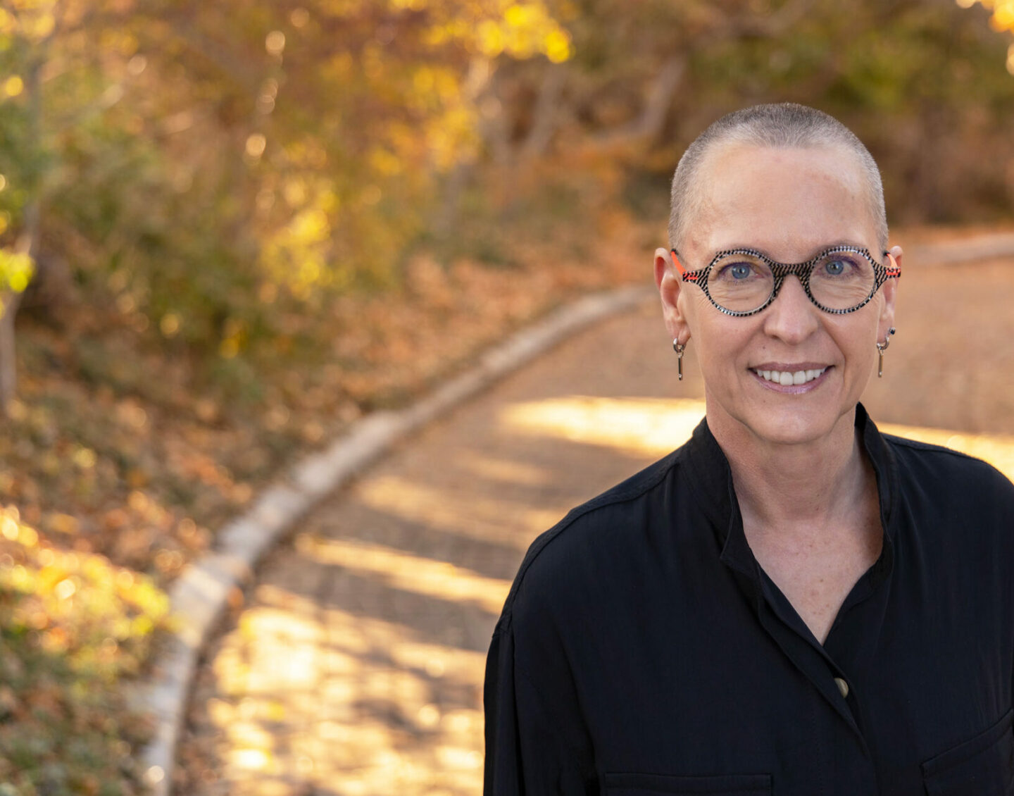 A white woman with a shaved head, glasses and a black shirt stands outdoors and smiles