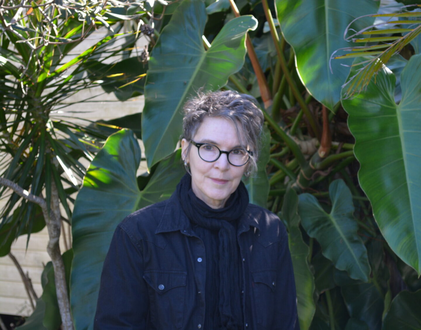 A white woman with round glasses and hair worn loosely up, standing in front of a plant with large leaves