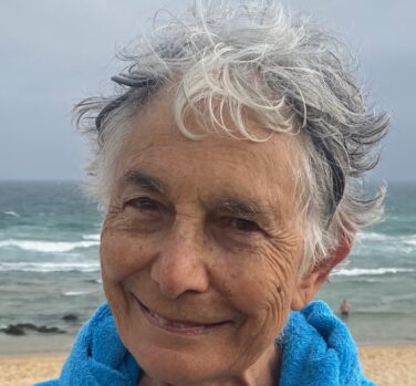 An older woman with short grey hair and a fleecy blue jacket stands smiling on a beach