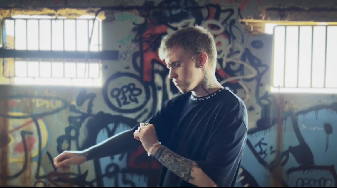 A man with short blonde hair, earring and sleeve tattoos dances, facing side-on to the camera