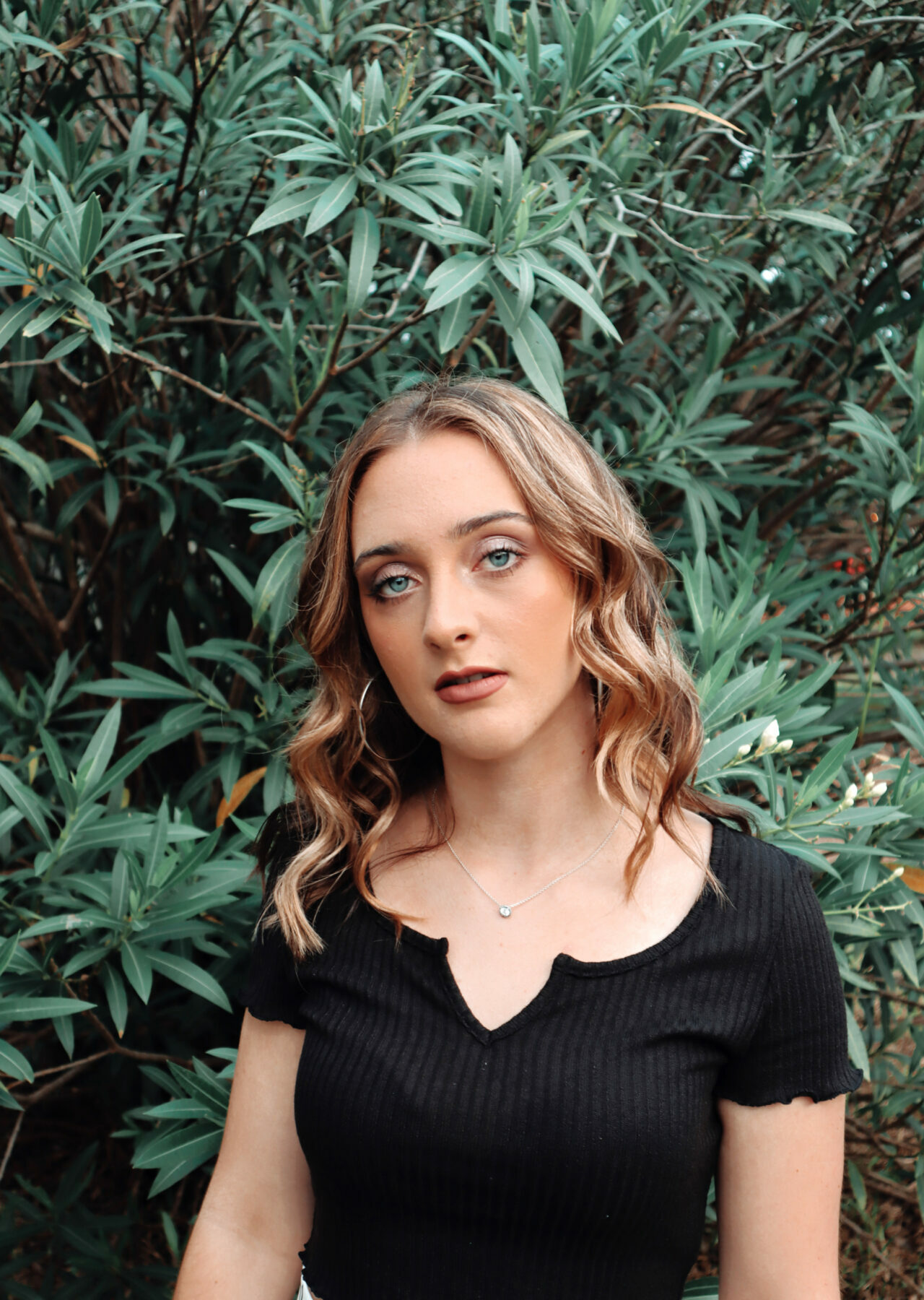 A young woman with wavy blonde hair stands in front of a green bush