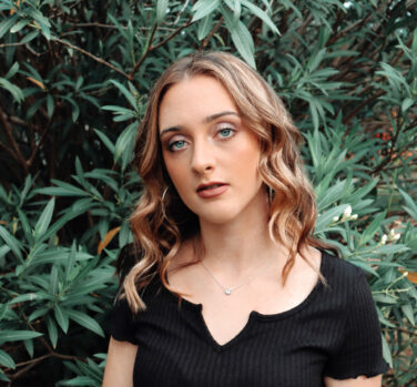 A young woman with wavy blonde hair stands in front of a green bush