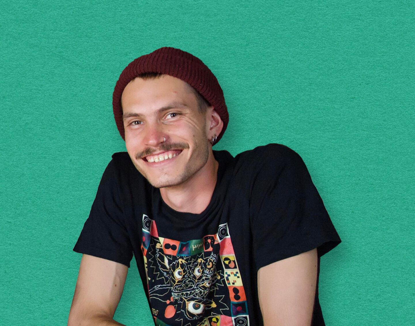 A young white man with a moustache, nose and ear piercings and red beanie leans over a yellow desk and smiles