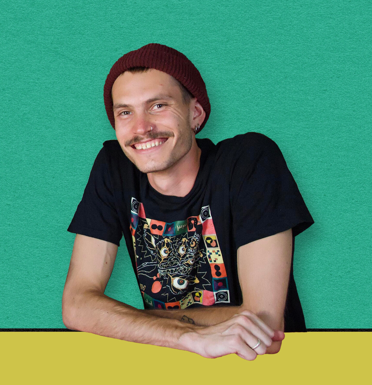 A young white man with a moustache, nose and ear piercings and red beanie leans over a yellow desk and smiles