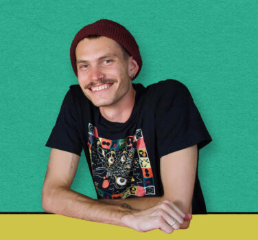 A young white man with a moustache, nose and ear piercings and red beanie leans over a yellow desk and smiles