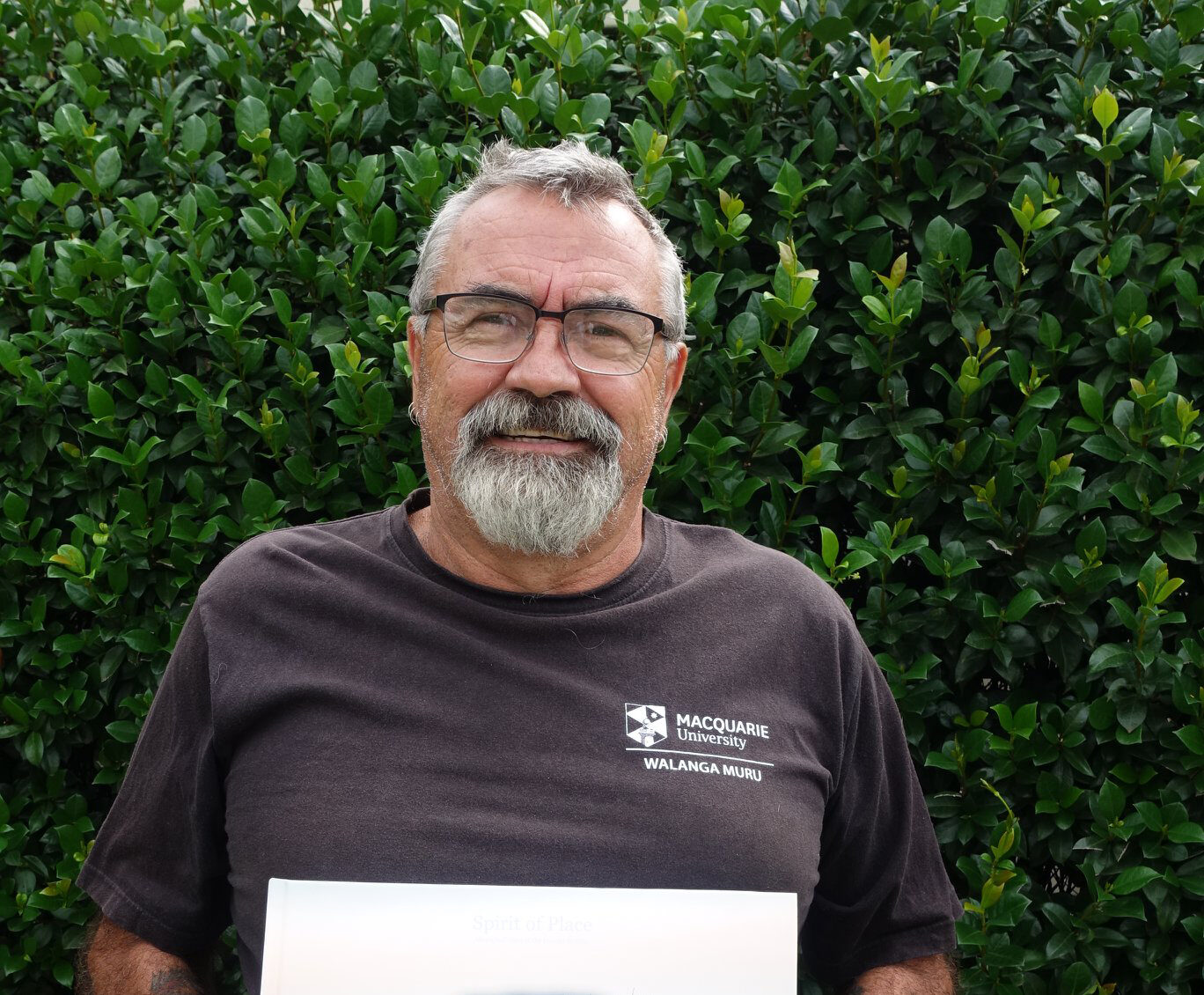 A man with glasses, short gray hair and circle beard stands outdoors holding a copy of his book and smiling.